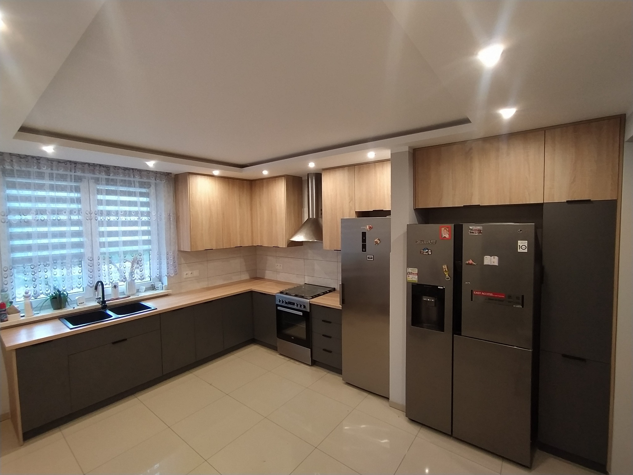 Nowoczesna kuchnia z drewnianymi górnymi szafkami i grafitowymi dolnymi, blatem z jasnego drewna i dużymi lodówkami side-by-side.