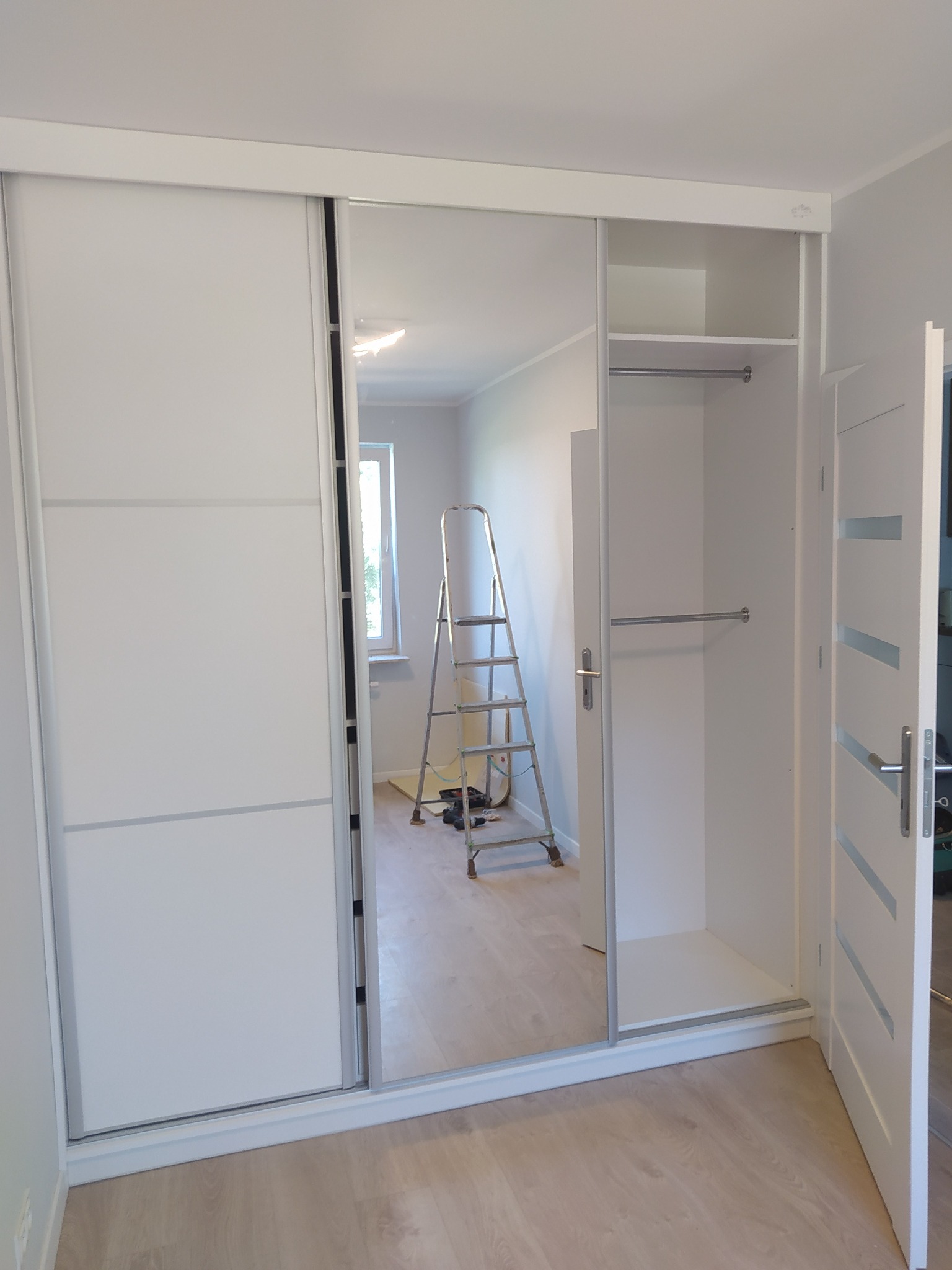 Biała szafa wnękowa z przesuwnymi drzwiami lustrzanymi, widoczne wnętrze z półkami i drążkami, drabina w tle, jasne panele podłogowe.
