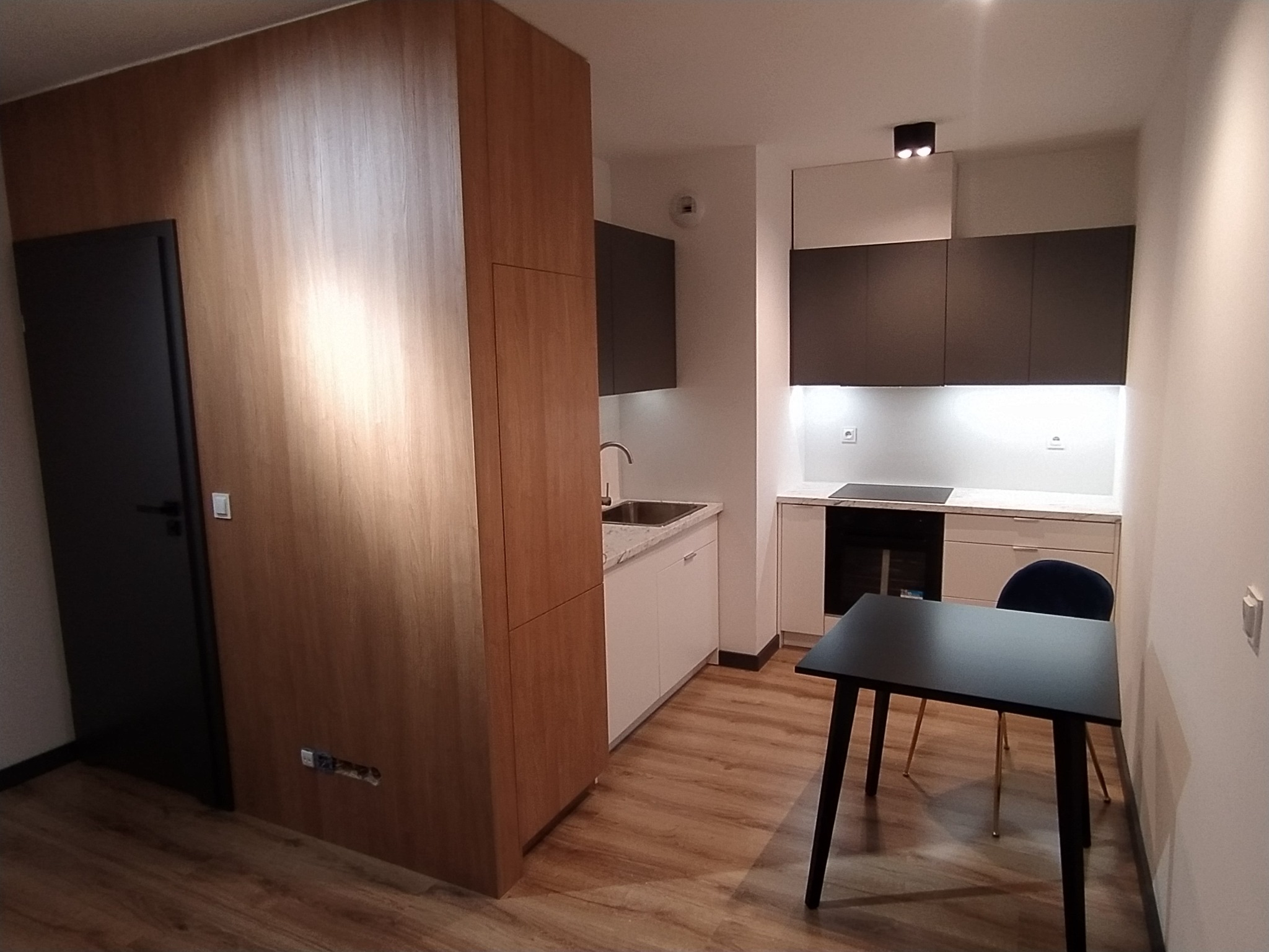 Nowoczesna kuchnia z zabudową w odcieniach brązu i szarości, z drewnianą szafą wnękową po lewej stronie i czarnym stołem z krzesłem na pierwszym planie.