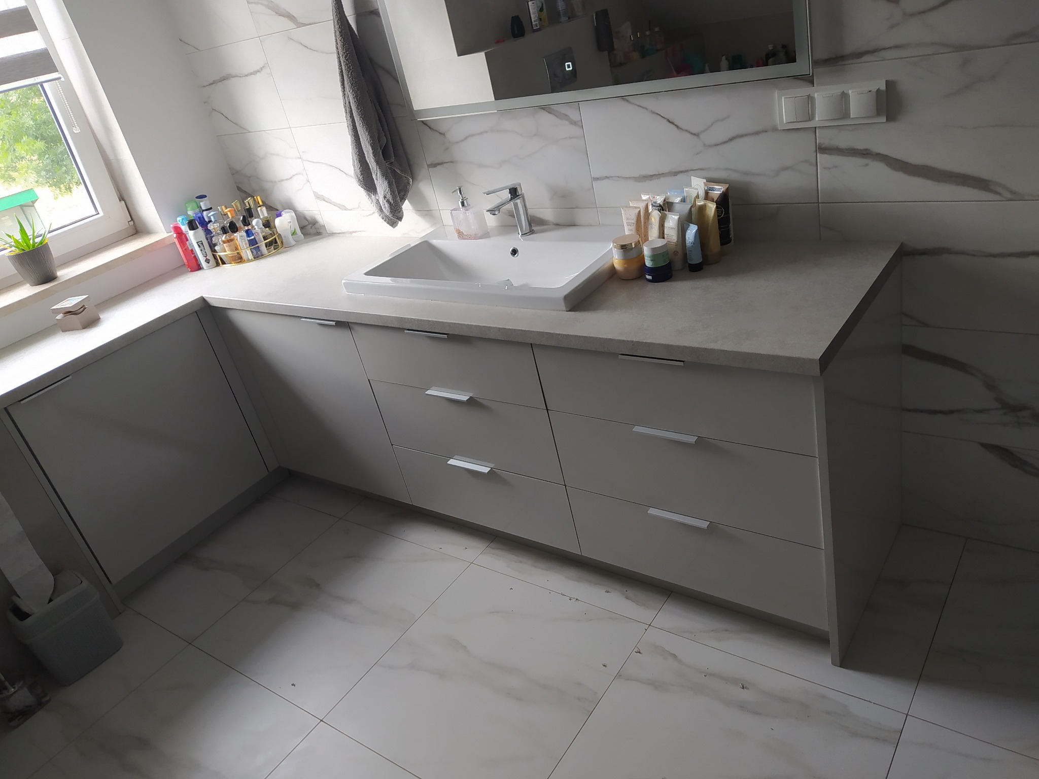 Jasna łazienka z szarymi szafkami pod umywalką, marmurowymi płytkami na ścianach i podłodze, oraz akcesoriami kosmetycznymi na blacie.