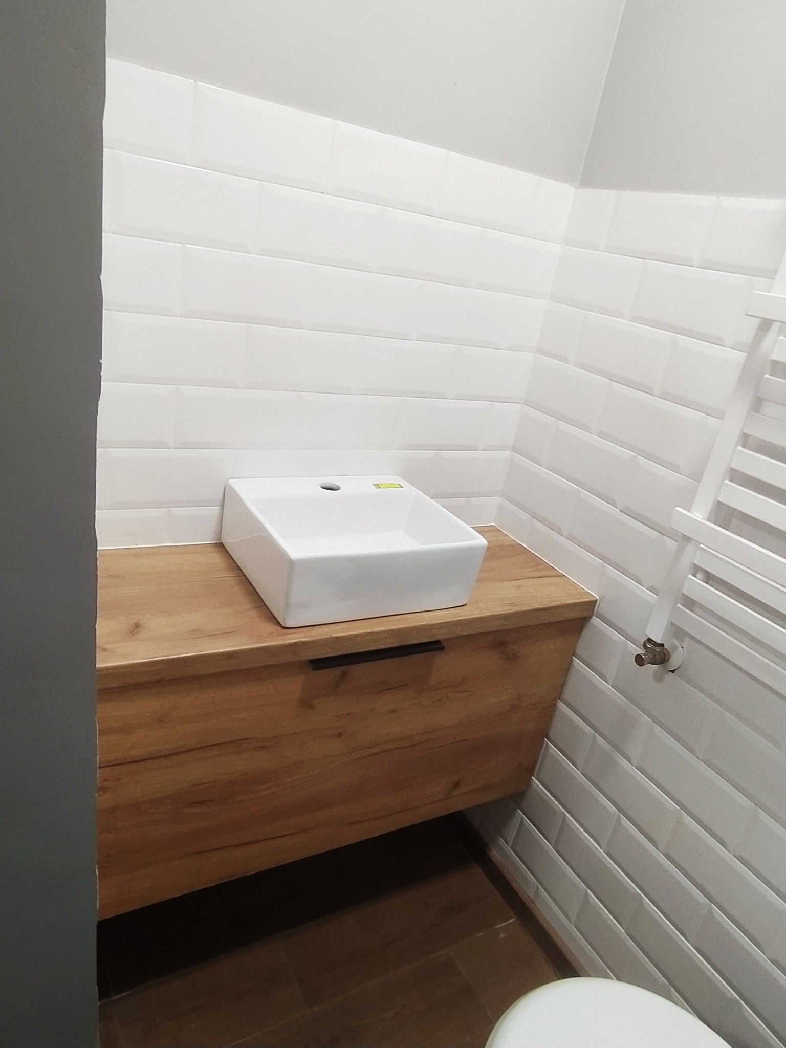 Nowoczesna łazienka z wiszącą szafką z drewnopodobnym blatem, na której umieszczona jest biała, prostokątna umywalka nablatowa. Białe, podłużne płytki na ścianach i grzejnik drabinkowy.