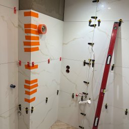 DomExpert Wrocław - Układanie płytek imitujących marmur w łazience, z użyciem poziomicy i klipsów. Widoczne fragmenty instalacji wodnej i taśma malarska na narożnikach.