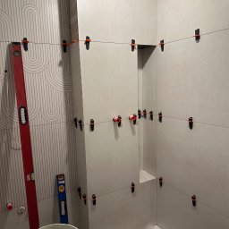 DomExpert Wrocław - Układanie płytek w łazience z użyciem poziomicy i klipsów dystansowych. Widoczny wzór na jednej ze ścian i wiadro z zaprawą. Prace w toku, etap przed fugowaniem.