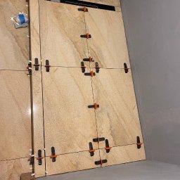 DomExpert Wrocław - Beżowe płytki imitujące marmur, ułożone na ścianie z użyciem systemu poziomowania płytek. Widoczny odpływ liniowy i szara ściana w tle.