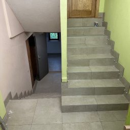 DomExpert Wrocław - Wykończone schody z szarych płytek, widok z góry. Na stopniach białe krzyżyki dystansowe, na dole poziomica i paca. Zielona ściana i drewniane drzwi w tle.