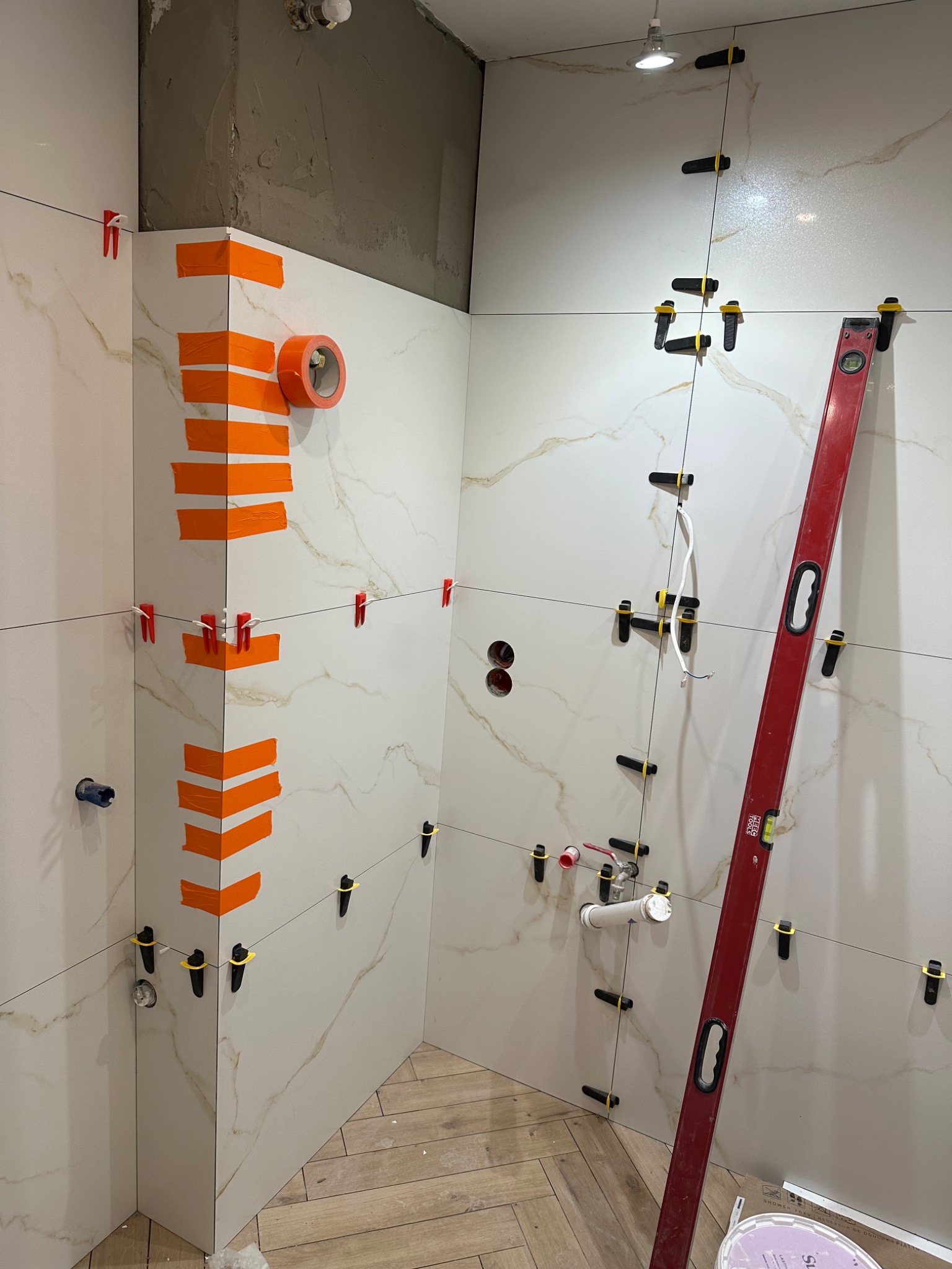 Układanie płytek imitujących marmur w łazience, z użyciem poziomicy i klipsów. Widoczne fragmenty instalacji wodnej i taśma malarska na narożnikach.