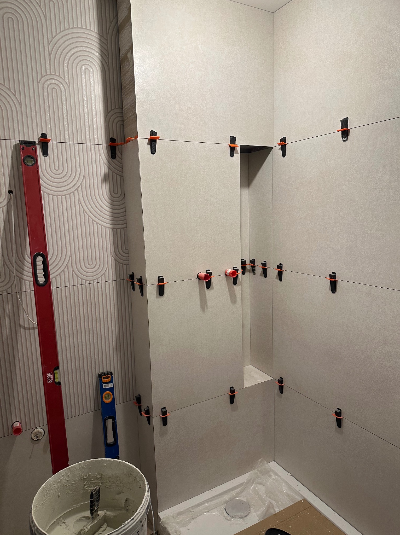 Układanie płytek w łazience z użyciem poziomicy i klipsów dystansowych. Widoczny wzór na jednej ze ścian i wiadro z zaprawą. Prace w toku, etap przed fugowaniem.