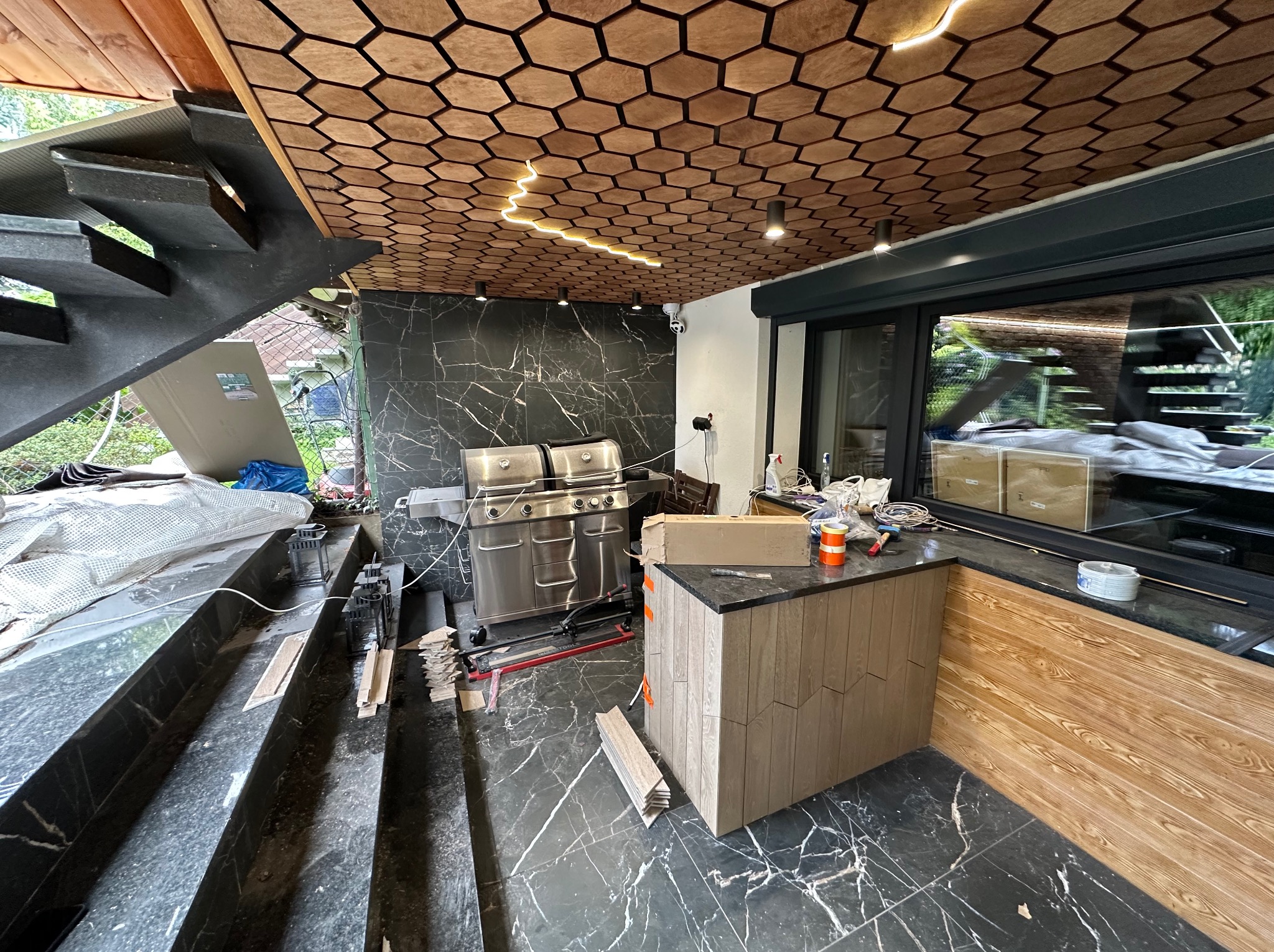 Wykończenie tarasu z marmurową podłogą i heksagonalnym sufitem z oświetleniem LED. Widoczny grill, fragment schodów i elementy wykończenia wnętrza.