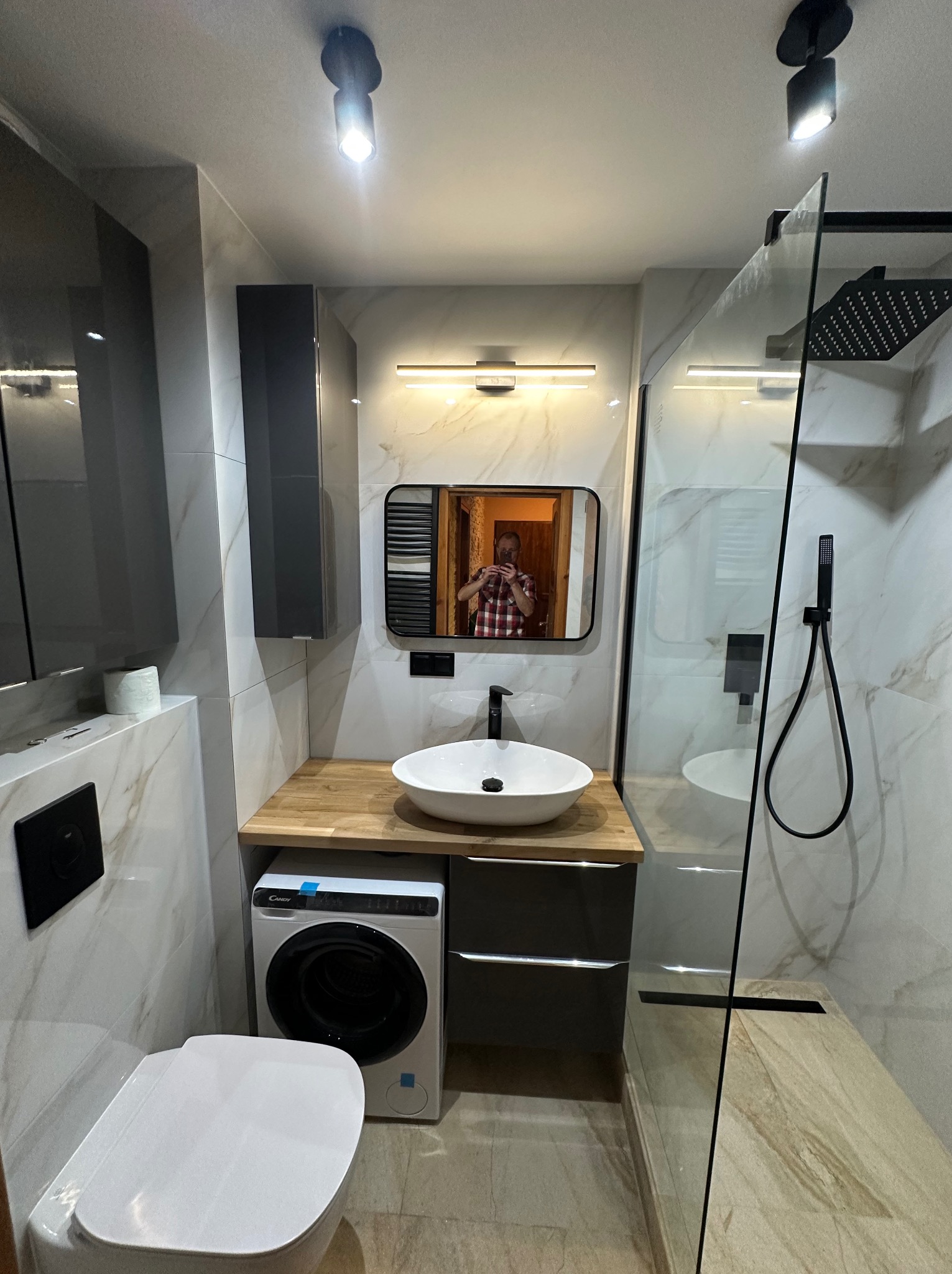 Nowoczesna łazienka z kabiną prysznicową typu walk-in, umywalką nablatową i pralką. Marmurowe płytki, czarne akcenty i oświetlenie punktowe.