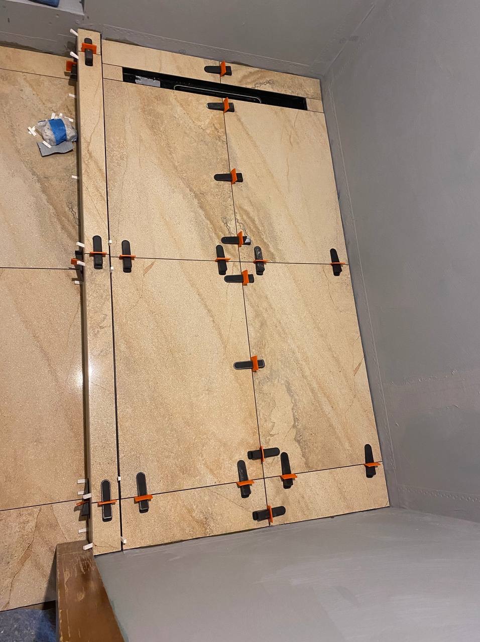 Beżowe płytki imitujące marmur, ułożone na ścianie z użyciem systemu poziomowania płytek. Widoczny odpływ liniowy i szara ściana w tle.