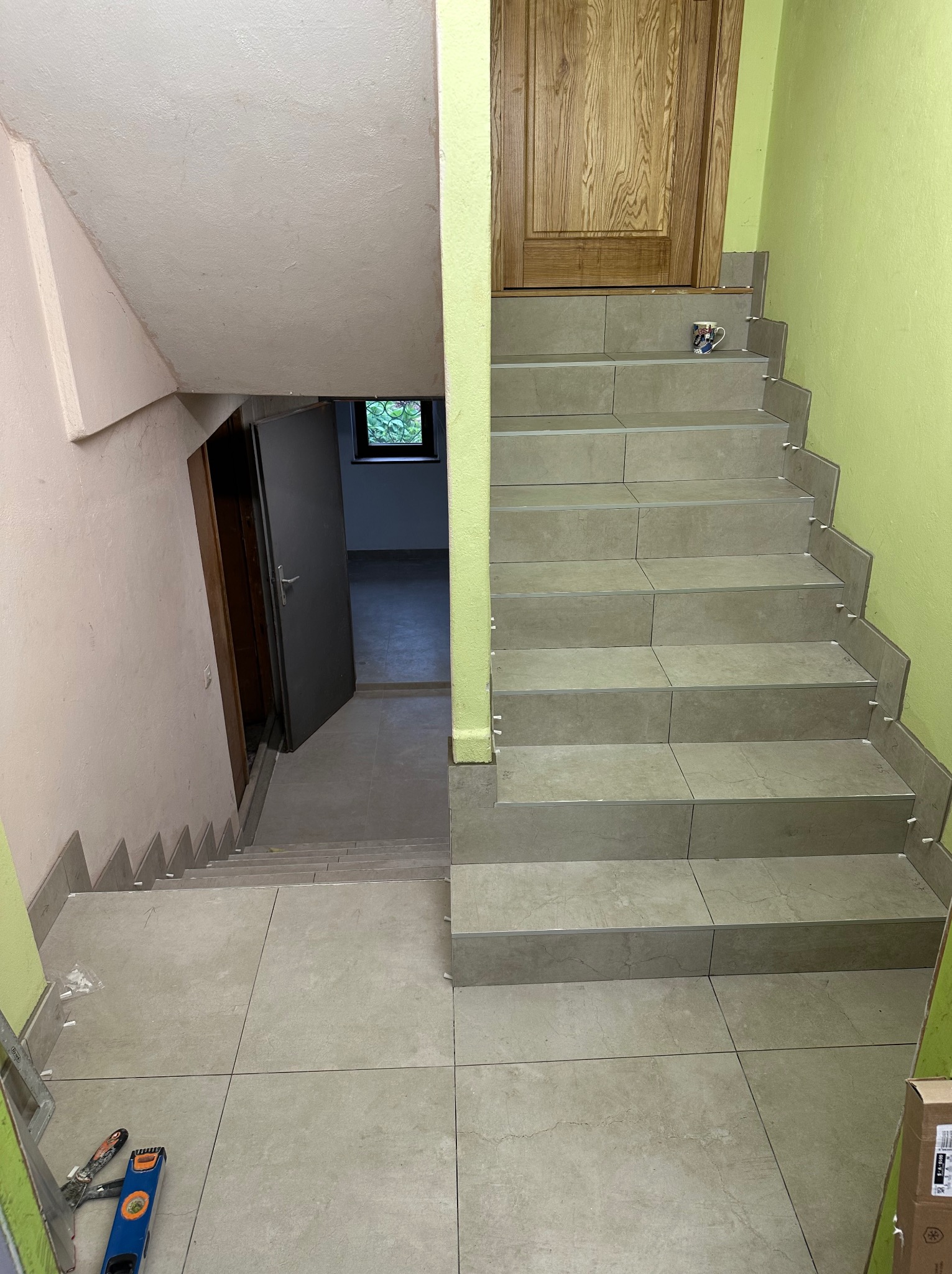 Wykończone schody z szarych płytek, widok z góry. Na stopniach białe krzyżyki dystansowe, na dole poziomica i paca. Zielona ściana i drewniane drzwi w tle.