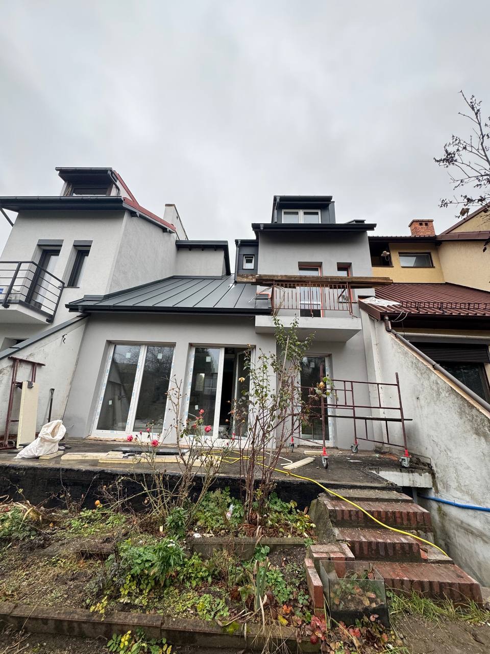 Elewacja domu w trakcie prac wykończeniowych: szare ściany, nowe okna, metalowy dach. Widoczne rusztowania i elementy ogrodu. Pochmurne niebo w tle.