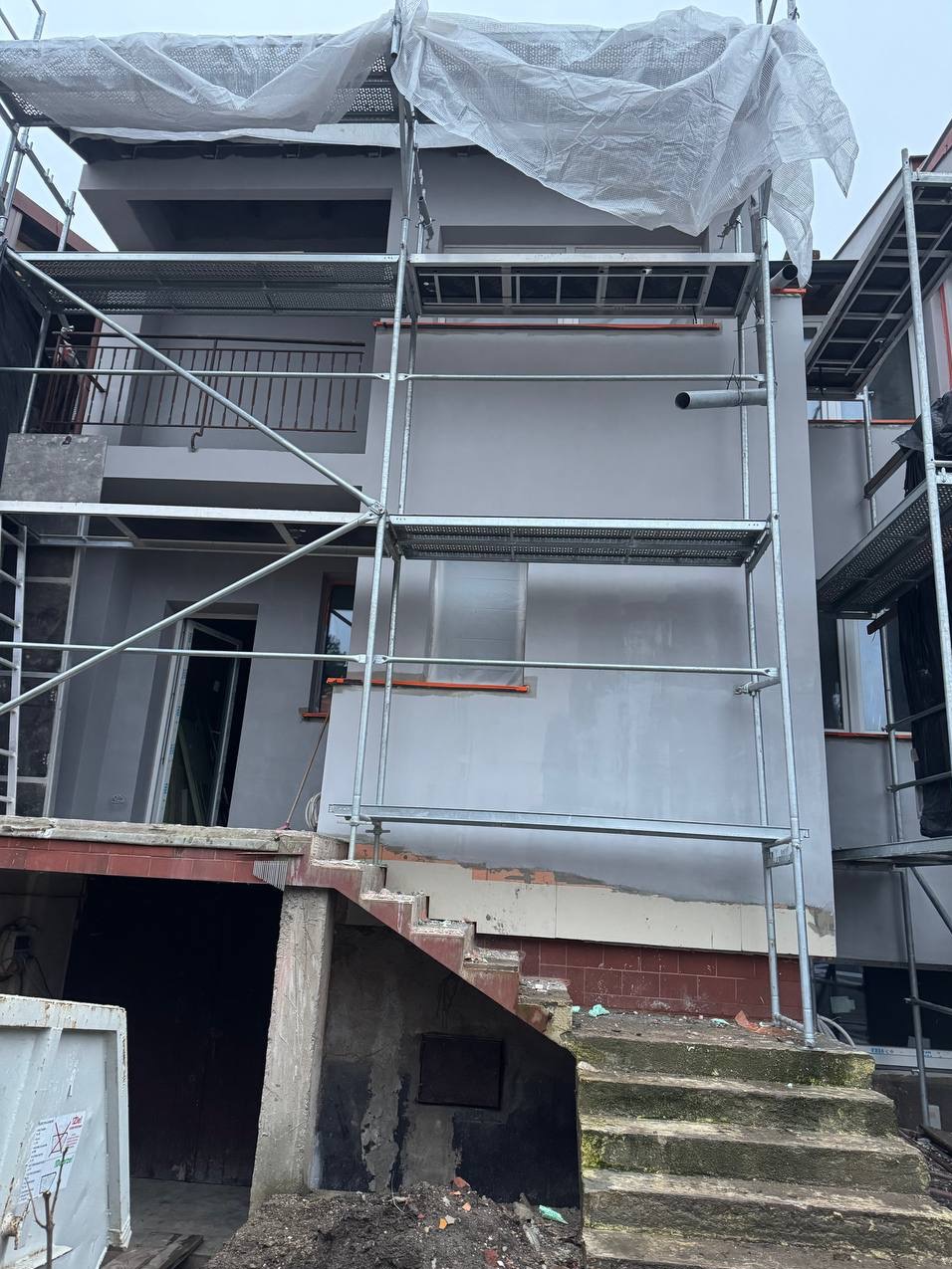 Budynek w trakcie prac elewacyjnych, widoczne rusztowania i fragmenty otynkowanej ściany. Częściowo odkryte fundamenty i schody wejściowe.