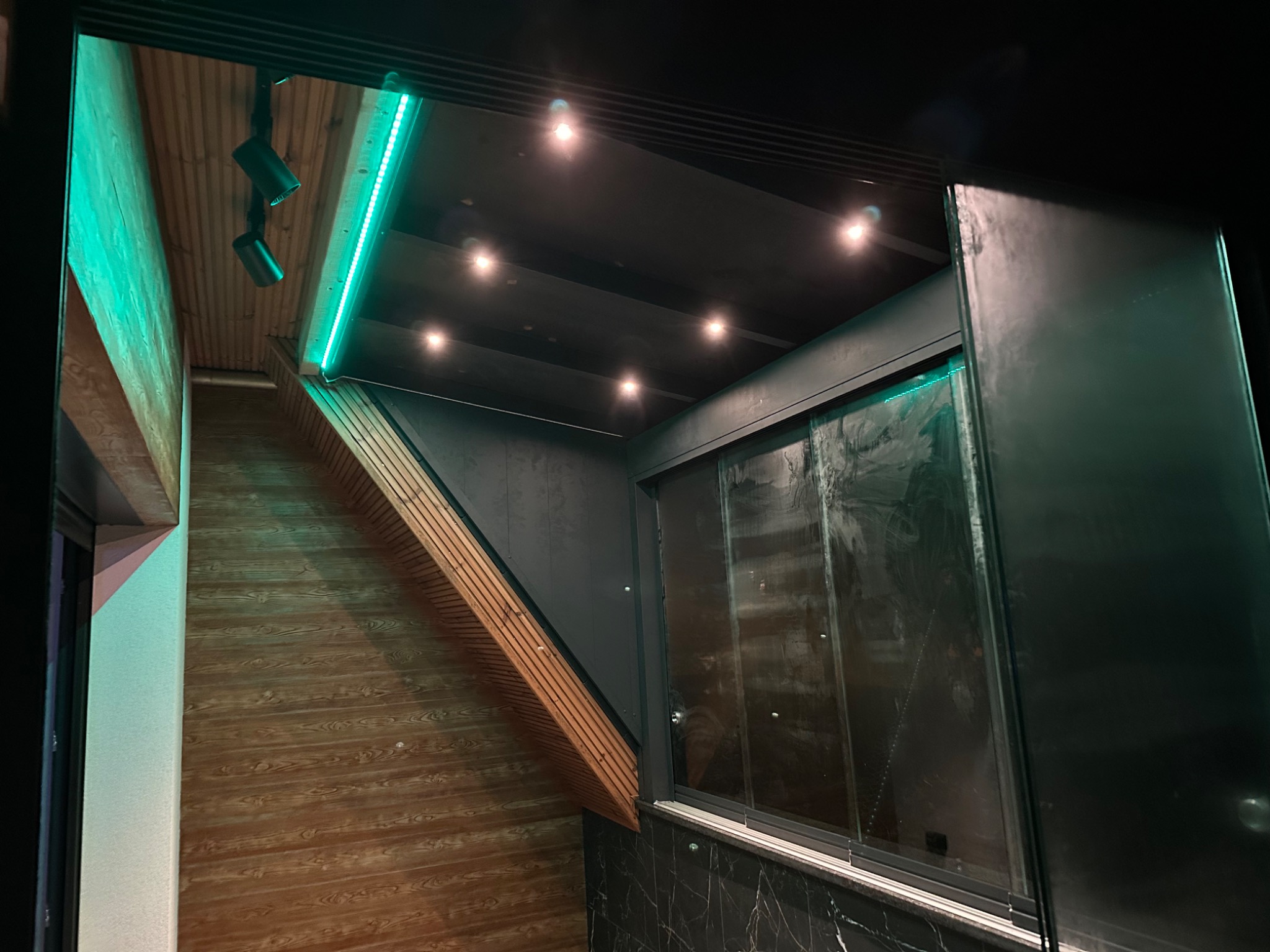 Nowoczesne wnętrze z drewnianą boazerią, oświetleniem LED i przesuwnymi szklanymi drzwiami. Czarny sufit z punktowymi lampami. Marmurowe wykończenie.