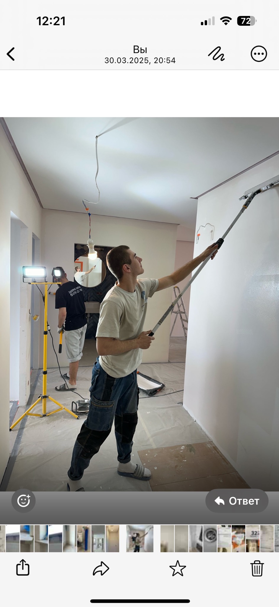 Mężczyzna w trakcie malowania ściany za pomocą wałka na długim kiju. W tle widoczny drugi pracownik i lampa oświetleniowa. Podłoga zabezpieczona folią.