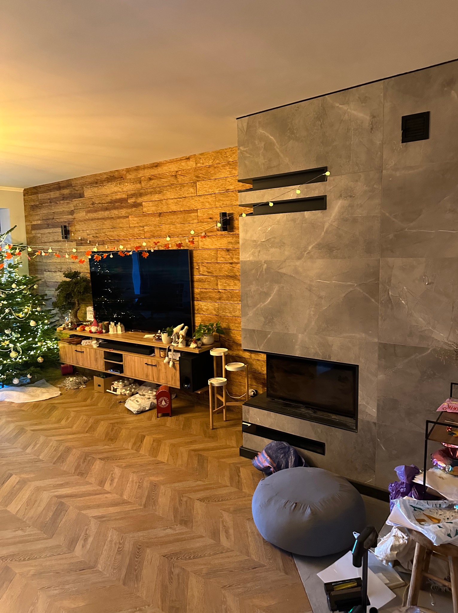 Salon z drewnianą ścianą, parkietem w jodełkę i kominkiem obłożonym szarymi płytami. Świąteczny wystrój z choinką i dekoracjami.