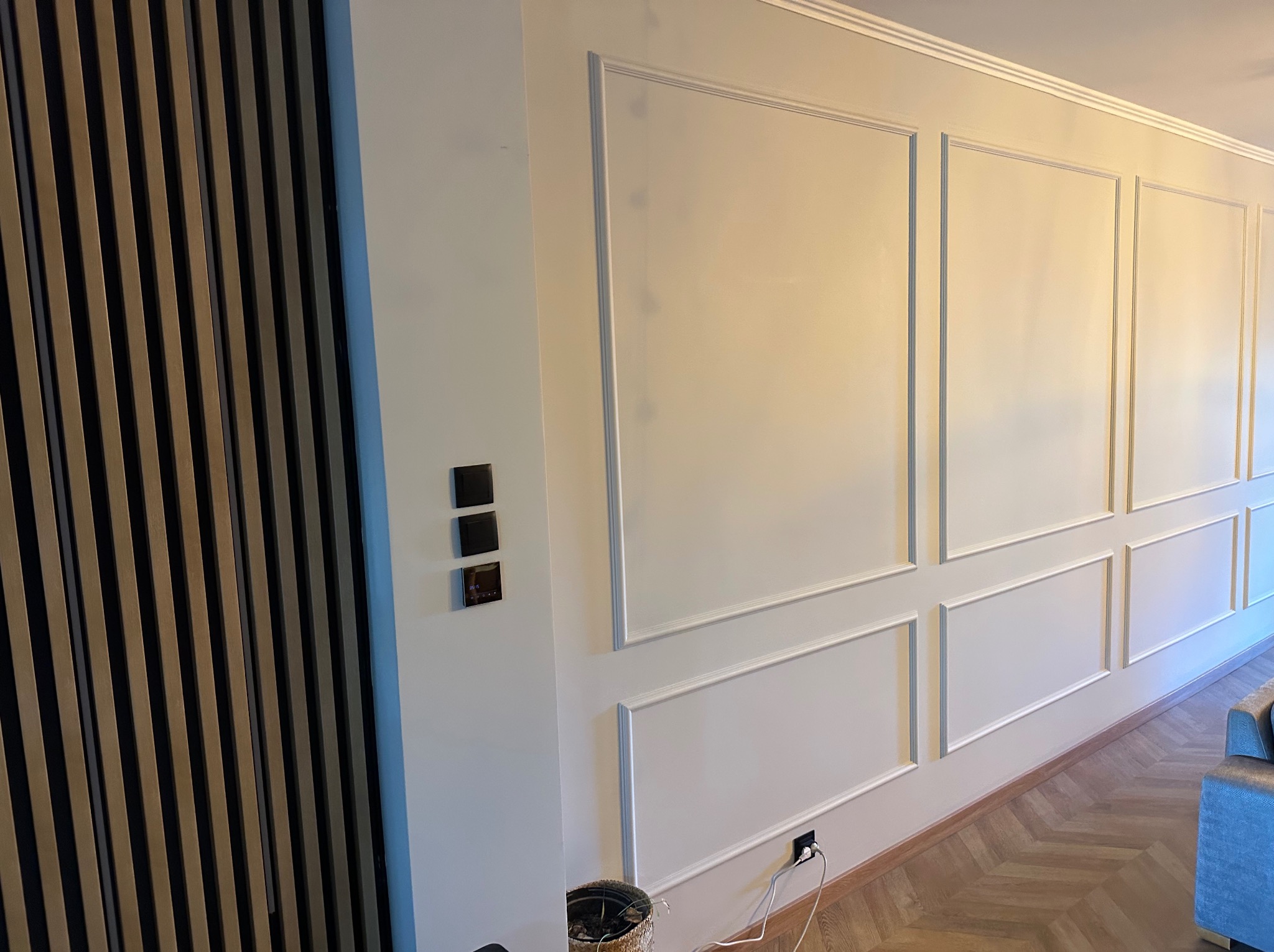 Eleganckie wykończenie ściany z dekoracyjnymi panelami i listwami w jasnym odcieniu, uzupełnione drewnianą okładziną i parkietem ułożonym w jodełkę.