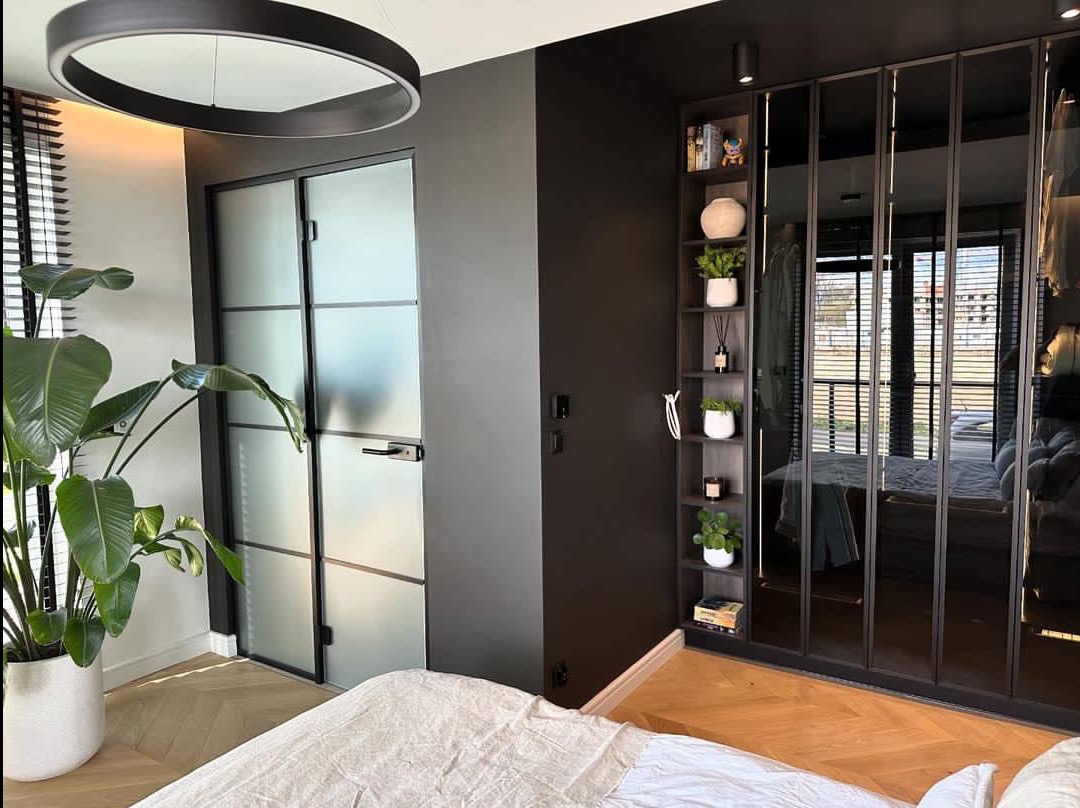 Nowoczesna sypialnia z ciemnymi ścianami, szklanymi drzwiami i dużą szafą z lustrami. Parkiet w jodełkę i okrągła lampa sufitowa dodają elegancji.