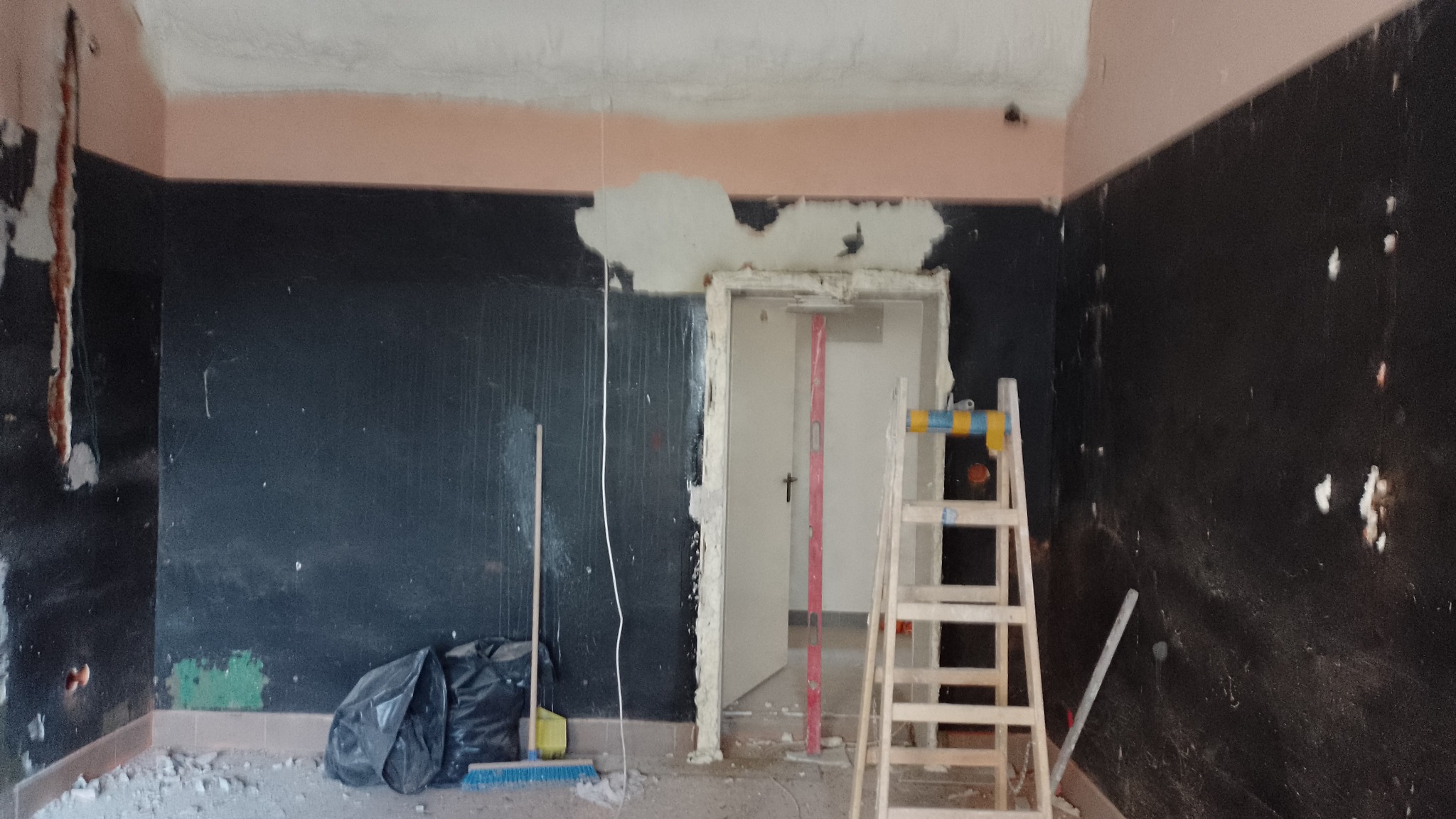 Pokój w trakcie remontu: czarne ściany z fragmentami starej farby, częściowo usunięta gładź przy suficie, drewniana drabina oparta o futrynę drzwiową, worki na gruz i narzędzia na podłodze.