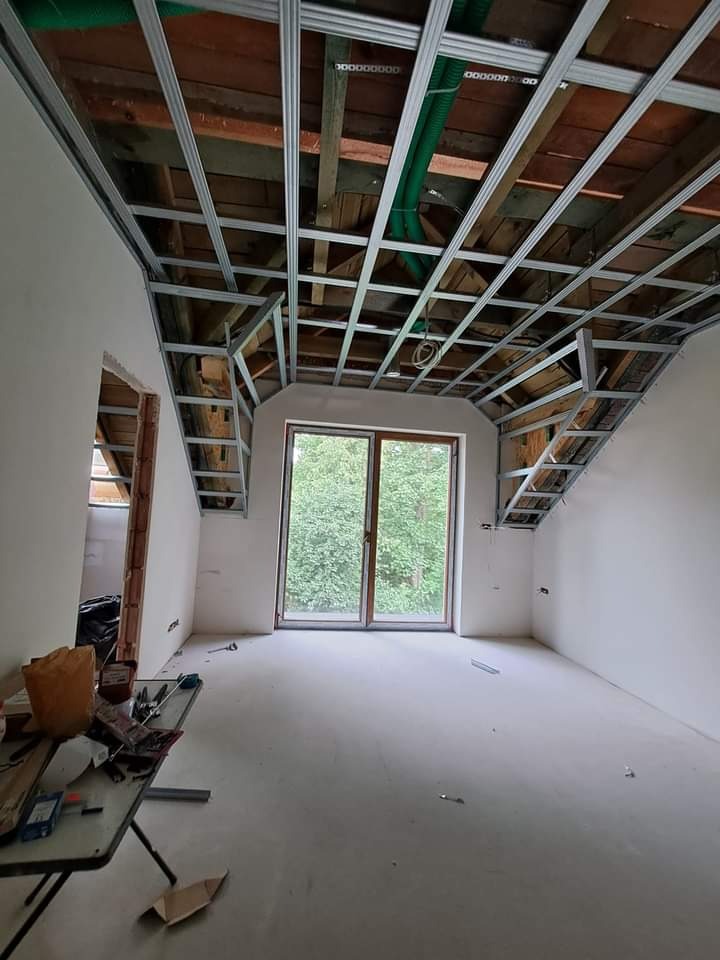 Poddasze w trakcie adaptacji: widok na stelaż sufitu podwieszanego z profili metalowych, przygotowany do montażu płyt gipsowo-kartonowych, z drewnianą konstrukcją dachu widoczną powyżej, okno...