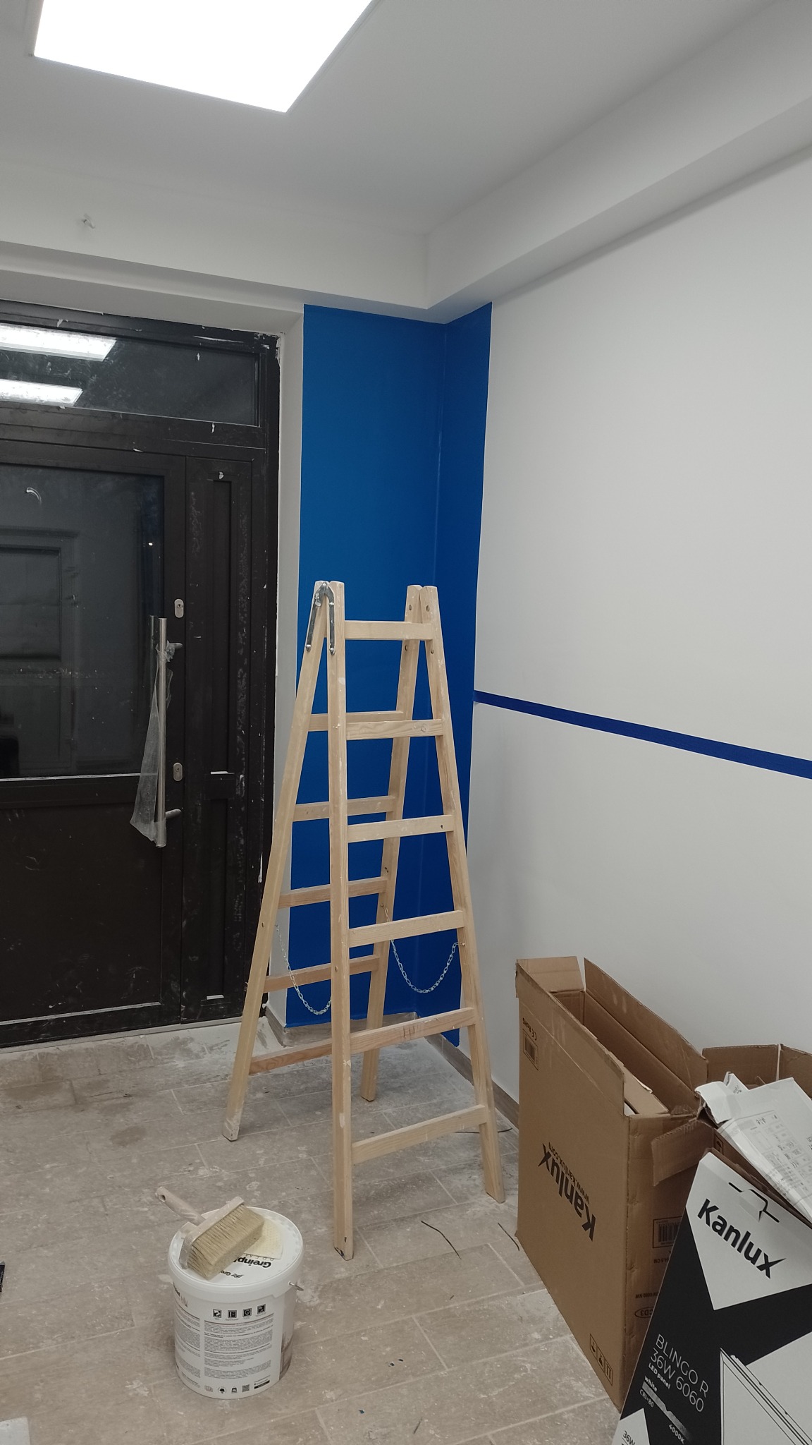 Wnętrze biura w trakcie odświeżania: ściana pomalowana na biało i intensywny niebieski, drewniana drabina, puszka farby z pędzlem obok i kartony po lampach Kanlux na podłodze.
