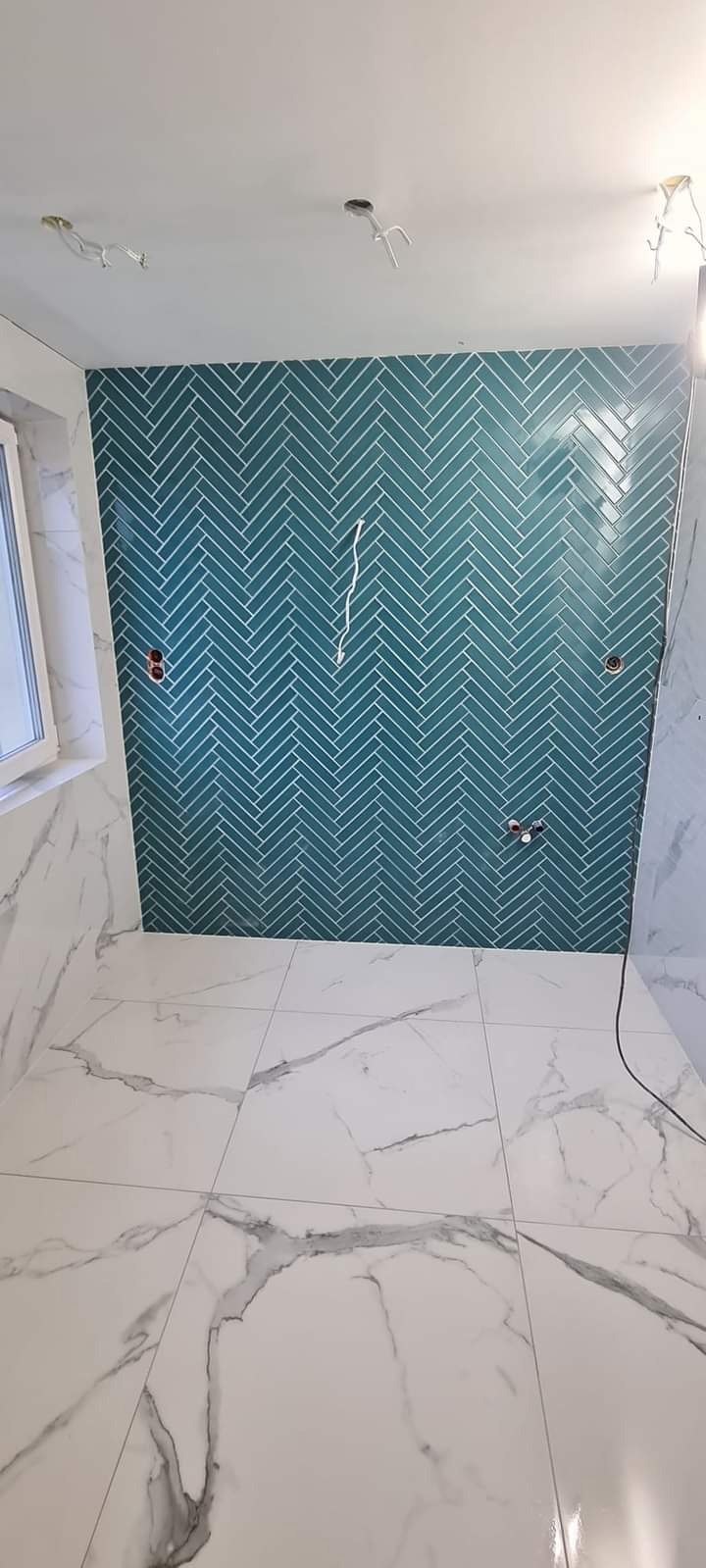 Wykończona łazienka z turkusową ścianą w jodełkę i marmurową podłogą, widoczne niedokończone punkty elektryczne i hydrauliczne.