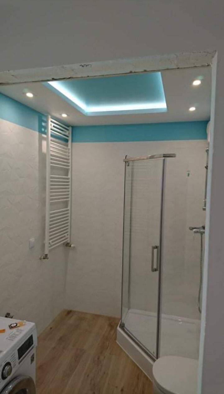 Wykończona łazienka z podświetlanym sufitem w kolorze turkusowym, kabiną prysznicową ze szklanymi drzwiami, grzejnikiem drabinkowym i pralką w rogu.