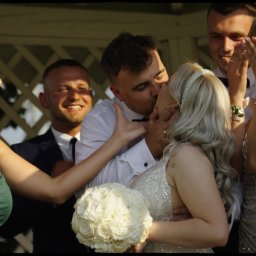 Od Serca Kamil Morawiec - Pocałunek nowożeńców w Modlnicy otoczonych rozbawionymi gośćmi weselnymi; panna młoda w sukni z cekinami trzyma bukiet białych róż.