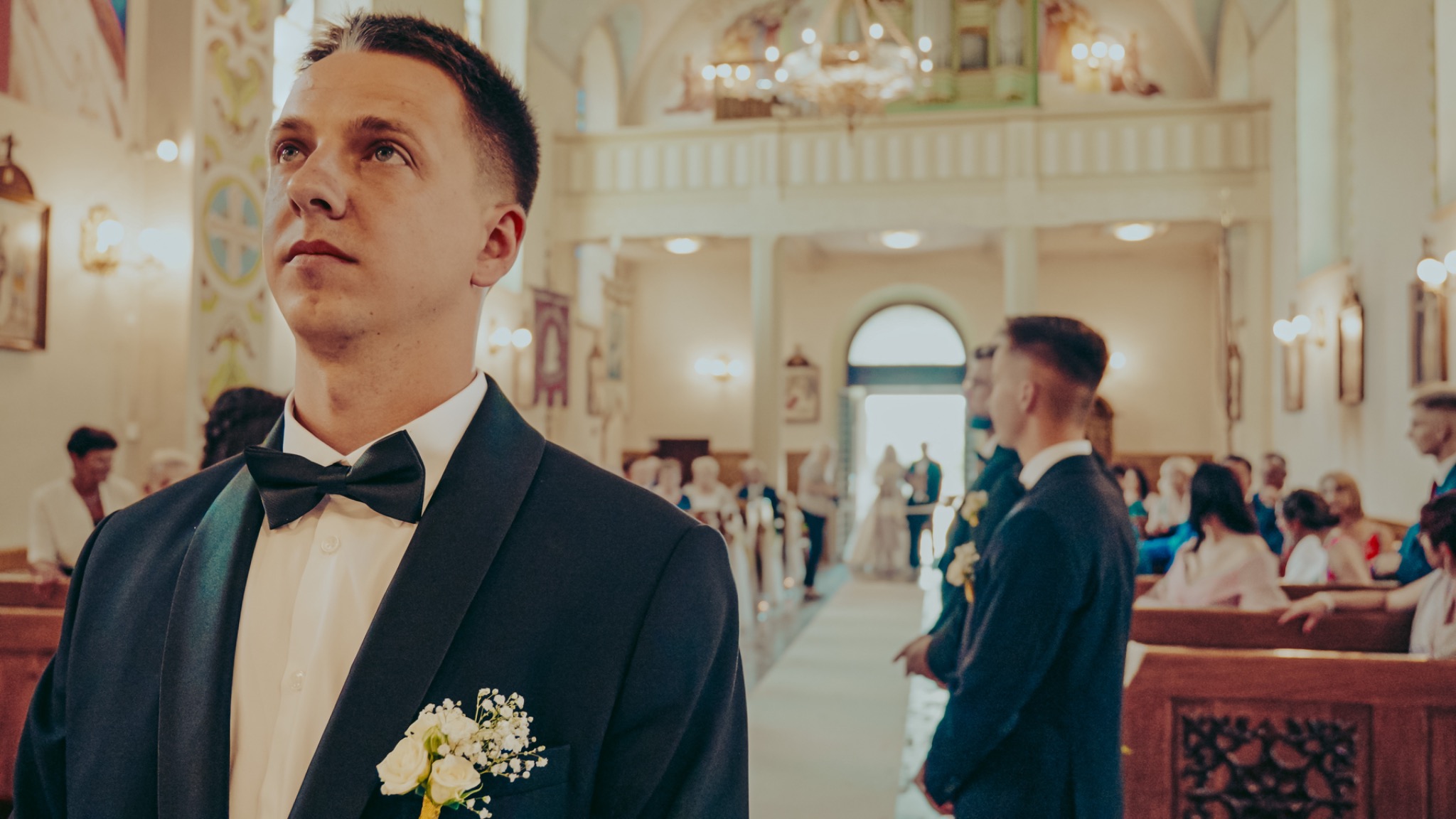 Pan młody w ciemnym smokingu z muchą i butonierką z białych kwiatów, oczekujący na ślub w kościele, widok z dołu.