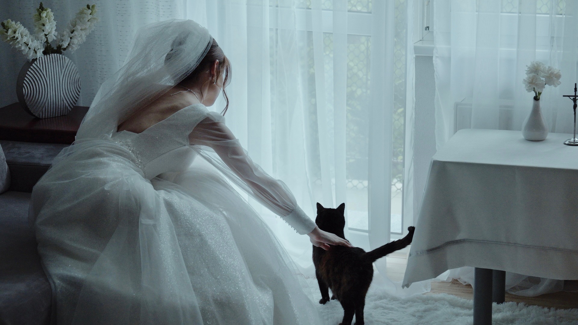 Panna młoda w białej sukni ślubnej z welonem siedzi na kanapie i głaszcze czarnego kota na tle okna z białymi firankami.