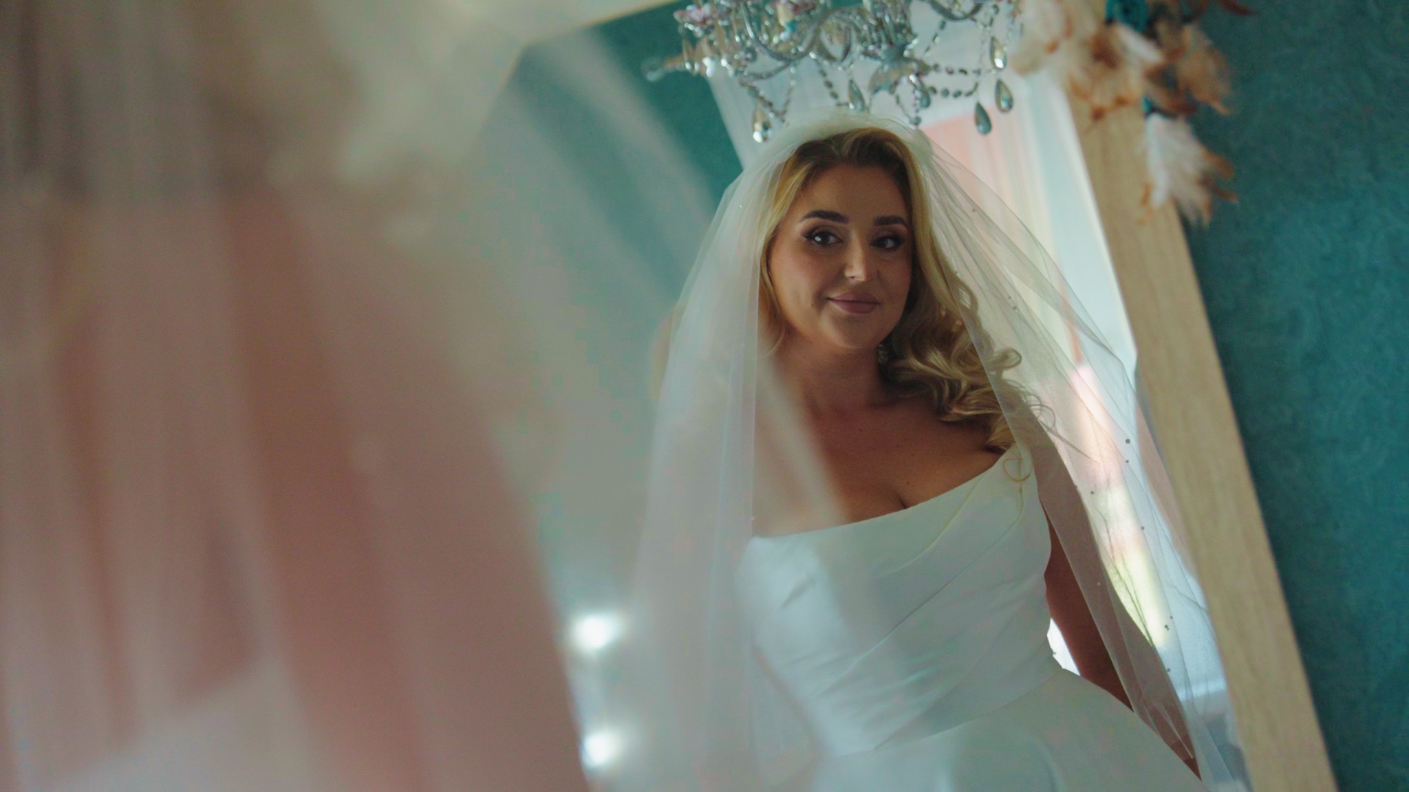 Panna młoda w białej sukni i welonie, spoglądająca w lustro, z kryształowym żyrandolem w tle.