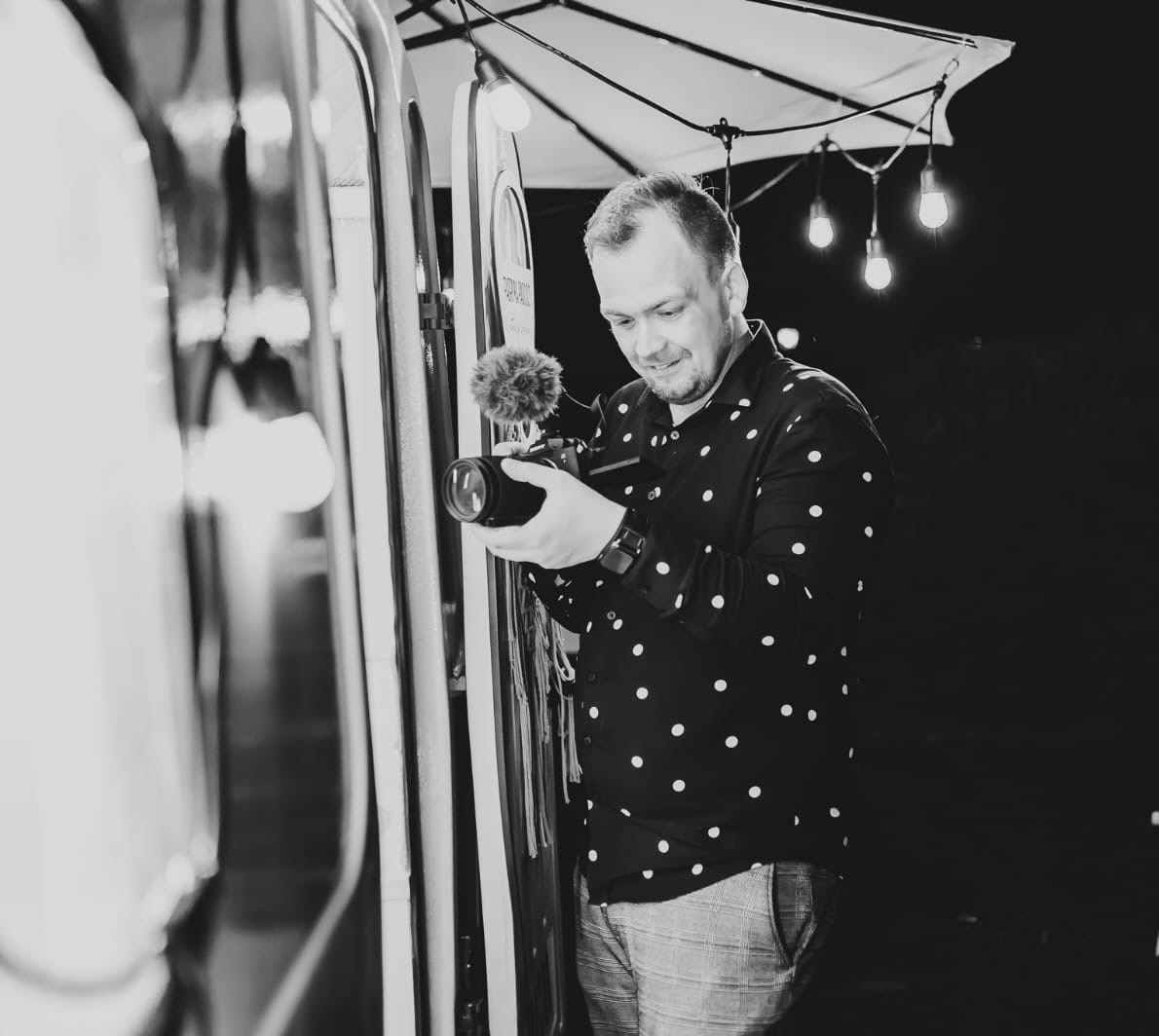 Kamerzysta w Modlnicy przegląda nagranie na ekranie aparatu z mikrofonem typu shotgun, stojąc obok oświetlonego wózka z akcesoriami.