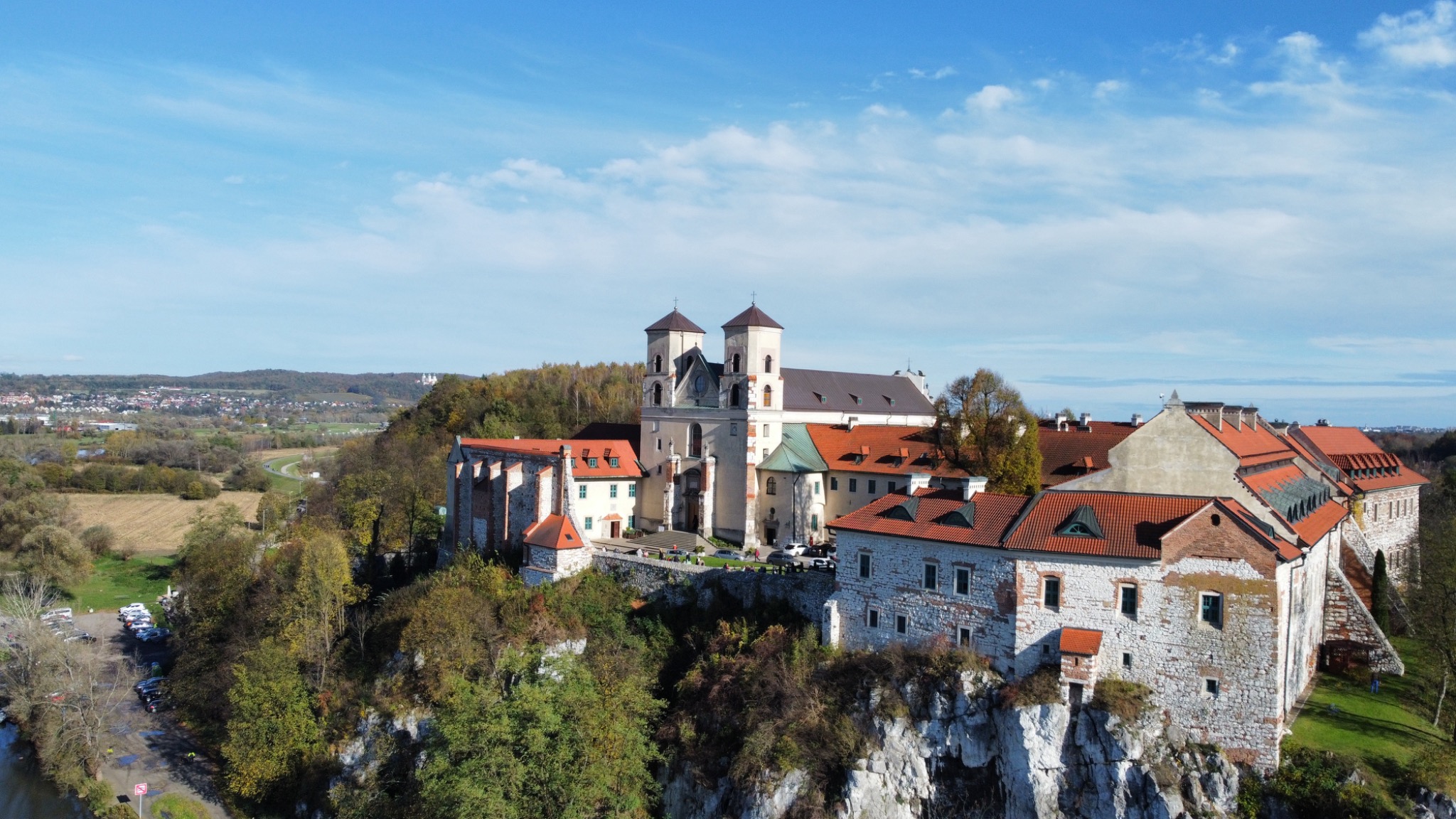 Opactwo Benedyktynów w Tyńcu, widok z lotu ptaka, budynki z czerwonymi dachami na skalistym wzgórzu, błękitne niebo z chmurami w tle.