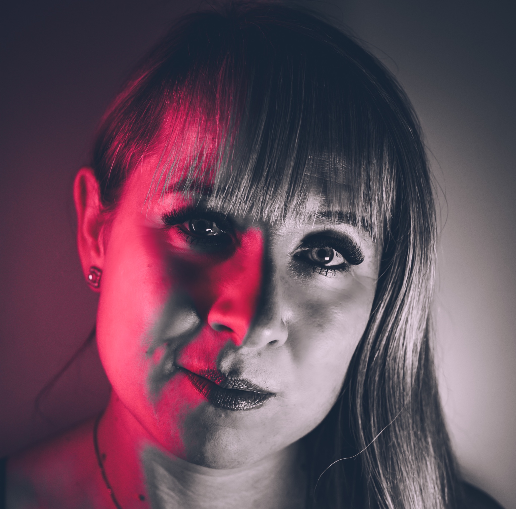 Portret kobiety z grzywką, oświetlonej czerwonym światłem po jednej stronie twarzy, z widocznym kolczykiem w uchu.