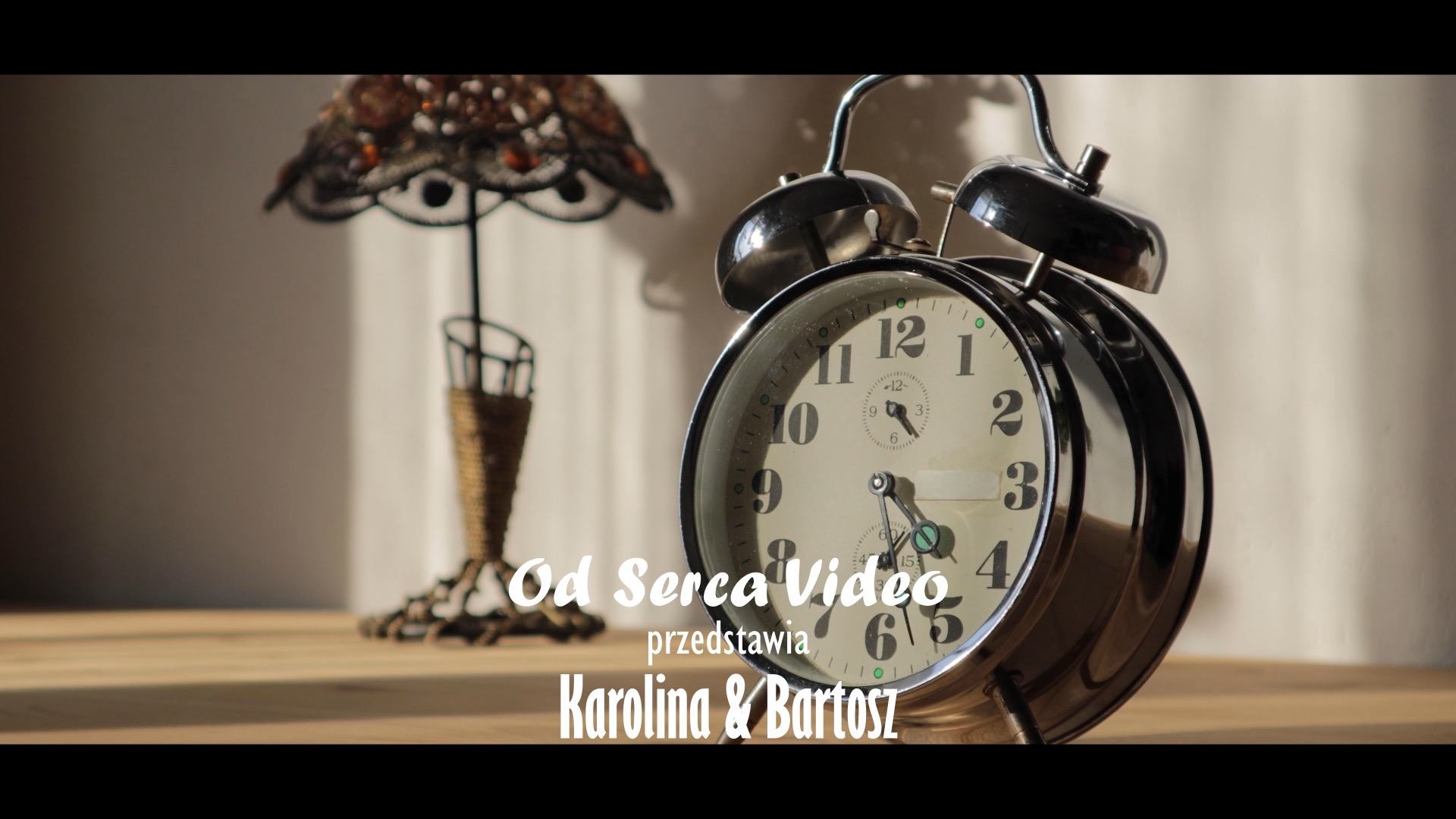 Stylizowany zegar budzik i lampka nocna na drewnianym stole, z nałożonym tekstem 'Od Serca Video przedstawia Karolina & Bartosz', sugerującym logo firmy.
