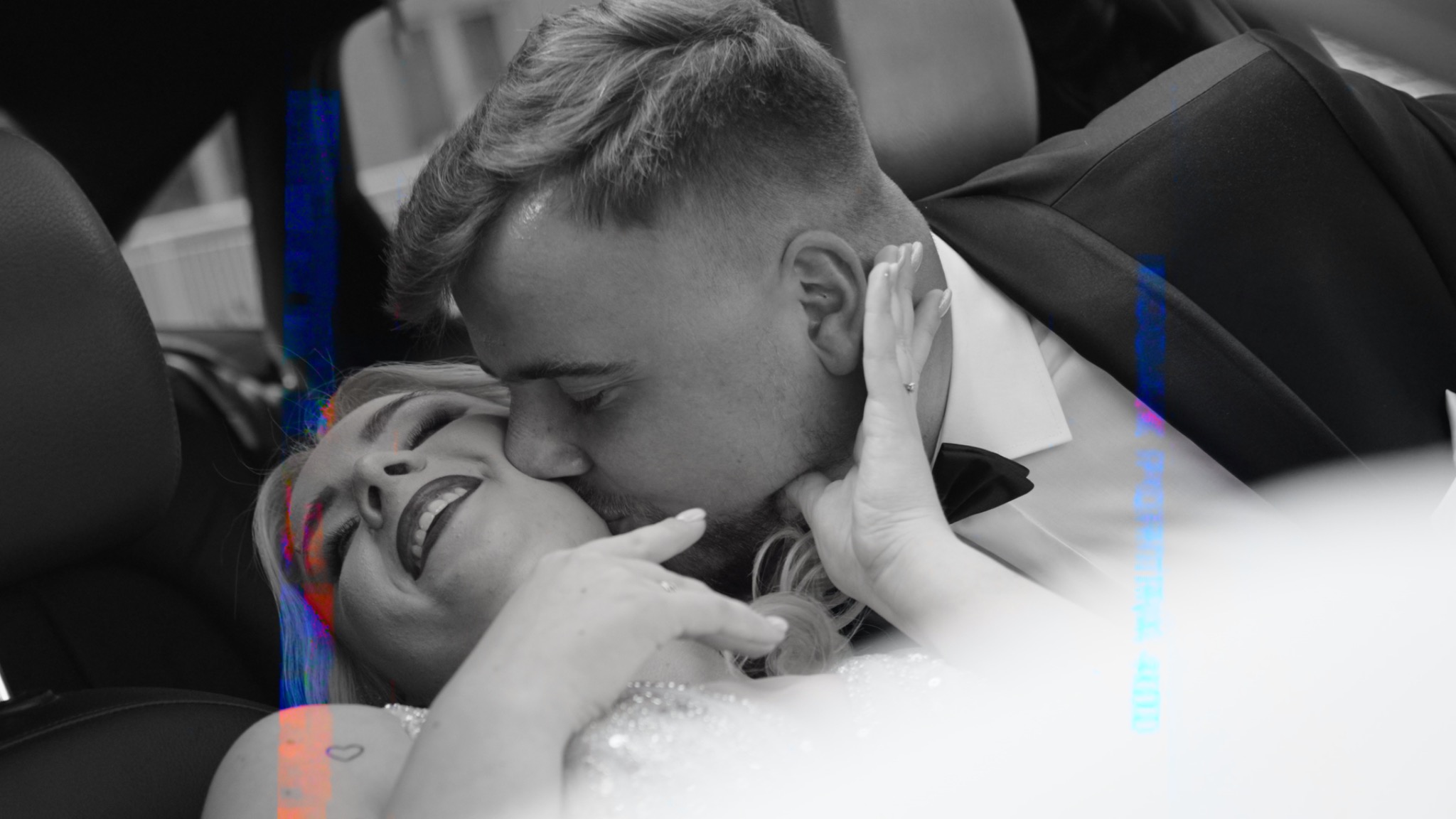 Czarno-białe ujęcie nowożeńców w samochodzie: Panna młoda z radością obejmuje twarz mężczyzny w smokingu, oboje w zbliżeniu, z delikatnym efektem wizualnym na zdjęciu.