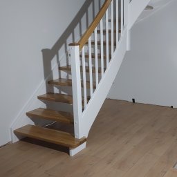 Mateusz Kowalczuk - Klatka schodowa z drewnianymi stopniami i białymi balustradami, widok na zakrzywiony bieg schodów w trakcie montażu parkietu na podłodze.