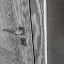 Mateusz Kowalczuk - Fragment drewnianych drzwi z widocznym usłojeniem w odcieniach szarości, metalową klamką i zamkiem, uchwycone z bliska pod kątem.