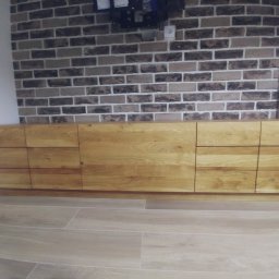 Mateusz Kowalczuk - Długa, niska szafka z jasnego drewna z widocznymi słojami, z szufladami i drzwiczkami, na tle ściany z cegły w odcieniach szarości i beżu, z widocznym gniazdkiem elektrycznym i elementami...