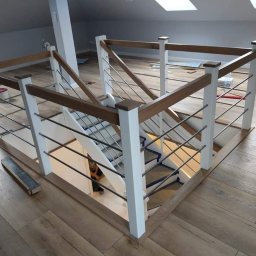 Mateusz Kowalczuk - Nowoczesne schody z białymi balustradami, drewnianymi poręczami i metalowymi prętami, widok z góry na klatkę schodową w jasnym wnętrzu poddasza z oknami dachowymi.