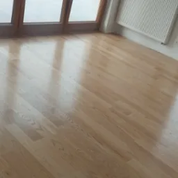 Jasny parkiet ułożony w nowym pokoju z dużymi drewnianymi oknami balkonowymi i białym grzejnikiem.