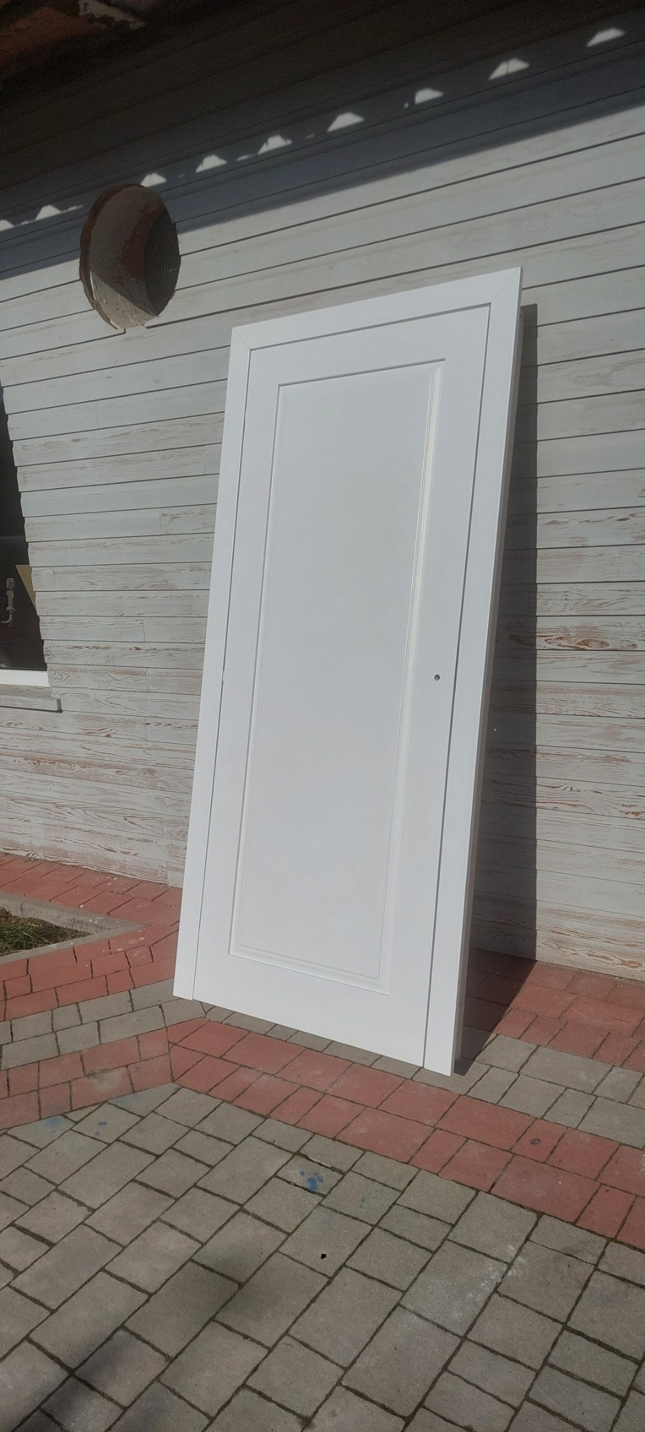 Białe, drewniane drzwi wewnętrzne o panelowej konstrukcji, oparte o elewację z desek i chodnik z kostki brukowej.