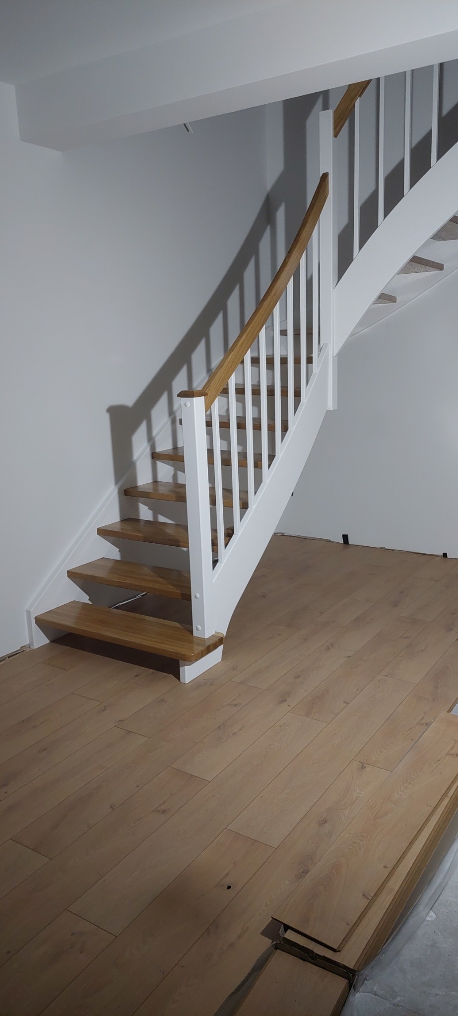 Klatka schodowa z drewnianymi stopniami i białymi balustradami, widok na zakrzywiony bieg schodów w trakcie montażu parkietu na podłodze.