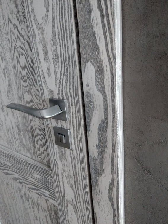 Fragment drewnianych drzwi z widocznym usłojeniem w odcieniach szarości, metalową klamką i zamkiem, uchwycone z bliska pod kątem.