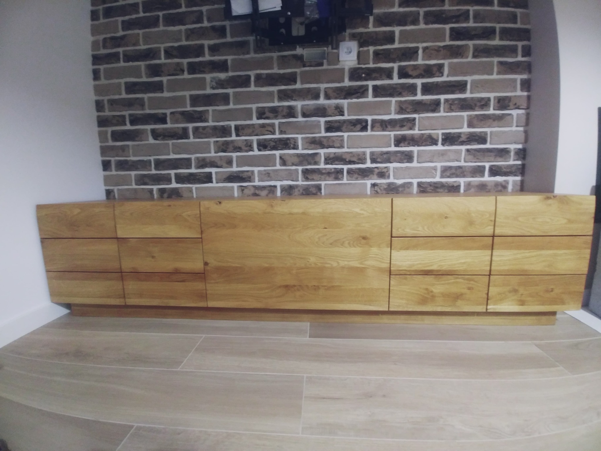 Długa, niska szafka z jasnego drewna z widocznymi słojami, z szufladami i drzwiczkami, na tle ściany z cegły w odcieniach szarości i beżu, z widocznym gniazdkiem elektrycznym i elementami...