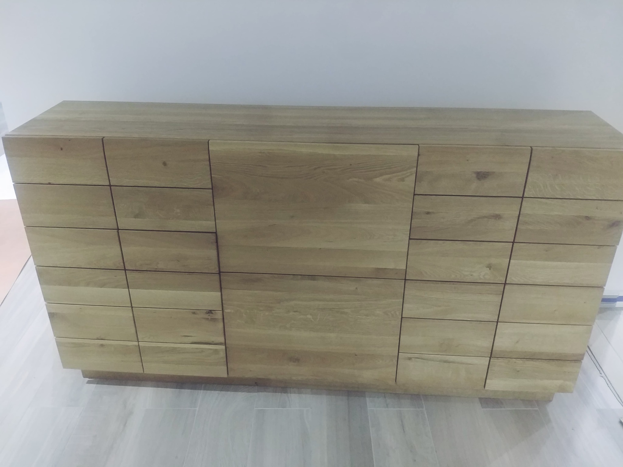 Nowoczesna szafka z jasnego drewna dębowego z frontami podzielonymi na prostokątne sekcje, umieszczona na podłodze wyłożonej płytkami imitującymi drewno.