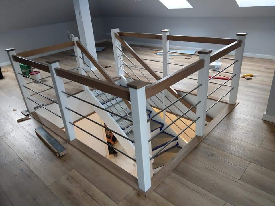 Nowoczesne schody z białymi balustradami, drewnianymi poręczami i metalowymi prętami, widok z góry na klatkę schodową w jasnym wnętrzu poddasza z oknami dachowymi.