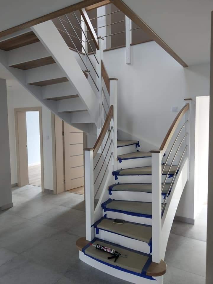 Nowoczesne schody z drewnianymi stopniami i białymi barierkami z metalowymi prętami, zabezpieczone niebieską taśmą malarską podczas prac wykończeniowych, tuba z klejem leży na jednym ze stopni.