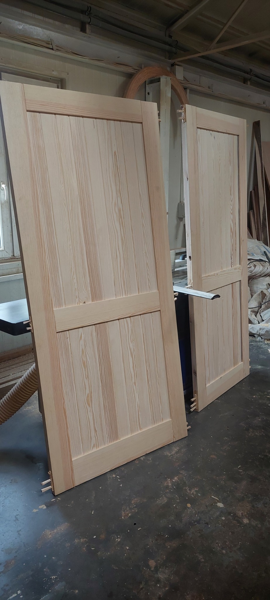 Dwa surowe skrzydła drzwiowe z jasnego drewna, z widocznym usłojeniem, o konstrukcji panelowej, ustawione w warsztacie stolarskim, z widocznymi kołkami montażowymi i fragmentem drewnianego łuku w tle.
