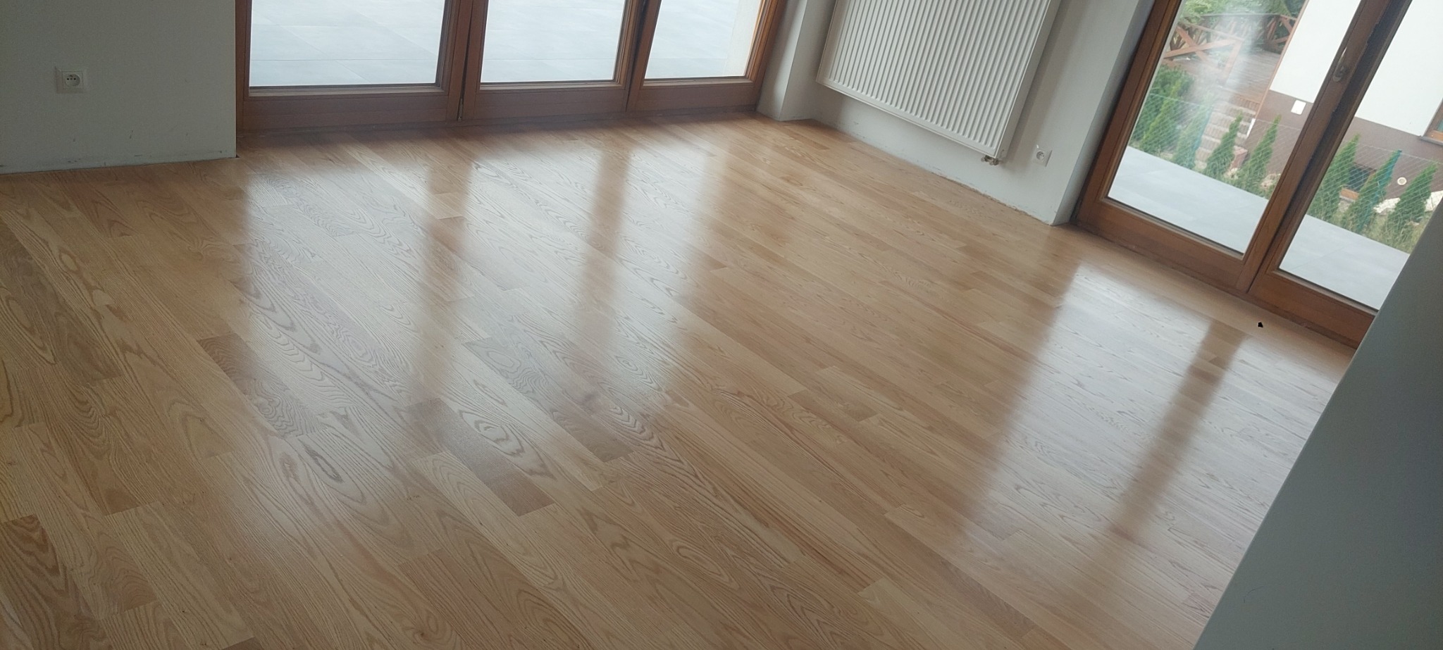Jasny parkiet ułożony w nowym pokoju z dużymi drewnianymi oknami balkonowymi i białym grzejnikiem.